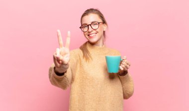 Genç sarışın kadın gülümsüyor ve arkadaş canlısı görünüyor. İki ya da iki numarayı gösteriyor. Elleri önde, geriye doğru sayıyor. kahve konsepti