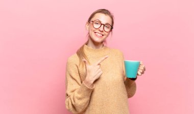 Genç sarışın kadın neşeli bir şekilde gülümsüyor, mutlu hissediyor ve kenarı ve yukarıyı işaret ediyor, fotokopi uzayında nesneler gösteriyor. kahve konsepti