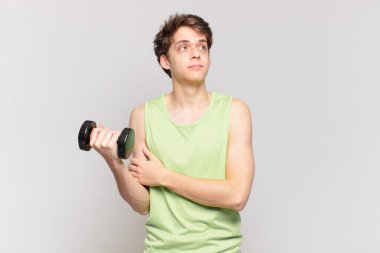 Genç bir çocuk omuz silkiyor, kafası karışık ve kararsız, kolları bağlı ve şaşkın görünüyor. dambıl konsepti