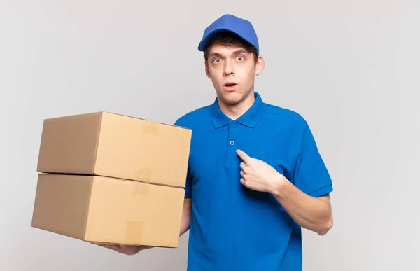 young package deliver boy looking shocked and surprised with mouth wide open, pointing to self