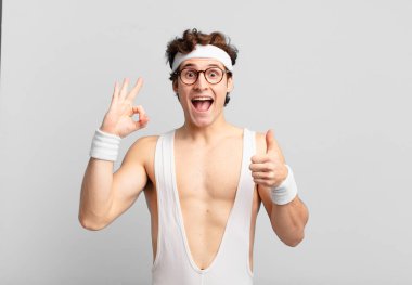 Komik sporcu kendini mutlu, şaşkın, tatmin olmuş ve şaşırmış hissediyor, tamam gösteriyor ve el kol hareketi yapıyor, gülümsüyor.