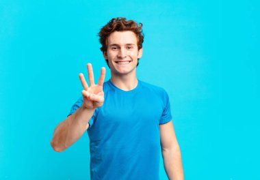 Genç sporcu çocuk gülümsüyor ve arkadaş canlısı görünüyor. Üç ya da üç numarayı gösteriyor. Elleri önde, geriye doğru sayıyor.