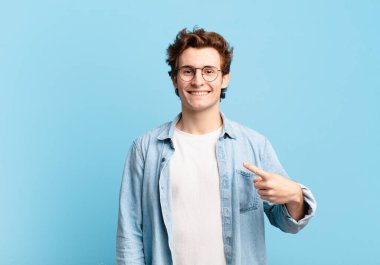 Genç yakışıklı çocuk neşeyle gülümsüyor, mutlu hissediyor ve kenarı ve yukarıyı işaret ediyor, fotokopi alanında nesneler gösteriyor.