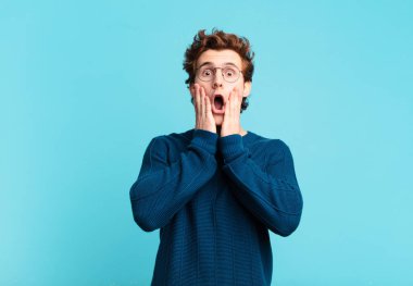 Genç yakışıklı çocuk şok olmuş ve korkmuş hissediyor, ağzı açık ve elleri yanaklarında korkmuş görünüyor.