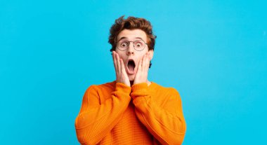 Genç yakışıklı çocuk şok olmuş ve korkmuş hissediyor, ağzı açık ve elleri yanaklarında korkmuş görünüyor.