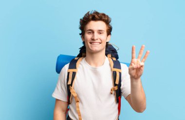 Yürüyüşçü çocuk gülümsüyor ve arkadaş canlısı görünüyor. Üç ya da üç numarayı gösteriyor. Elleri önde, geri sayıyor.