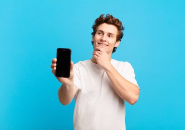 Genç çocuk mutlu bir şekilde gülümsüyor ve hayal kuruyor ya da şüphe ediyor, yan tarafa bakıyor. telefon ekranı kavramı