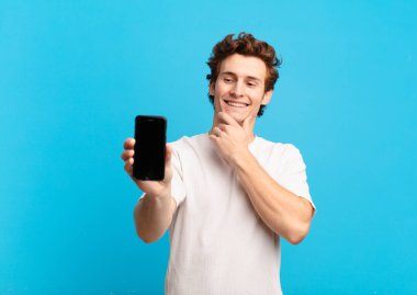 Genç bir çocuk mutlu, kendinden emin bir ifadeyle gülümsüyor, çenesi elleri çenesinde, merak ediyor ve yana bakıyor. telefon ekranı kavramı