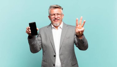 Kıdemli iş adamı gülümsüyor ve arkadaş canlısı görünüyor. Üç ya da üç numarayı gösteriyor. Elleri önde, geriye doğru sayıyor.