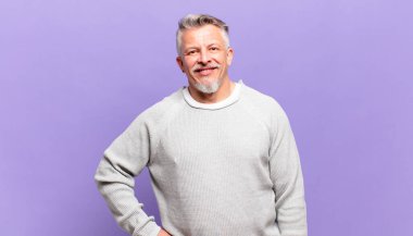 Yaşlı adam mutlu bir şekilde gülümsüyor, eli kalçasında ve kendinden emin, olumlu, gururlu ve arkadaş canlısı bir tavırla.