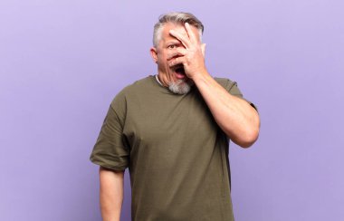 old senior man looking shocked, scared or terrified, covering face with hand and peeking between fingers
