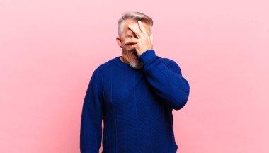 old senior man looking shocked, scared or terrified, covering face with hand and peeking between fingers