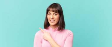 Güzel bir kadın neşeyle gülümsüyor, mutlu hissediyor ve kenarı ve yukarıyı işaret ediyor, fotokopi uzayındaki nesneleri gösteriyor.