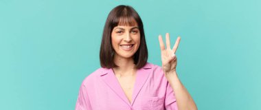 Güzel bir kadın gülümsüyor ve arkadaş canlısı görünüyor. Üç ya da üç numarayı gösteriyor. Elleri önde, geri sayıyor.