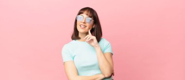 Güzel bir kadın mutlu, kendinden emin bir ifadeyle gülümsüyor, çenesinde elleri, merak ediyor ve yan tarafa bakıyor.