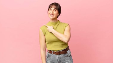 Güzel bir kadın neşeyle gülümsüyor, mutlu hissediyor ve kenarı ve yukarıyı işaret ediyor, fotokopi uzayındaki nesneleri gösteriyor.