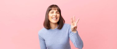 Güzel bir kadın gülümsüyor ve arkadaş canlısı görünüyor. Üç ya da üç numarayı gösteriyor. Elleri önde, geri sayıyor.