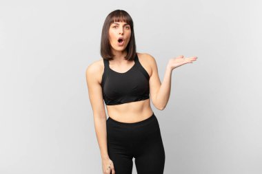 athlete woman looking surprised and shocked, with jaw dropped holding an object with an open hand on the side