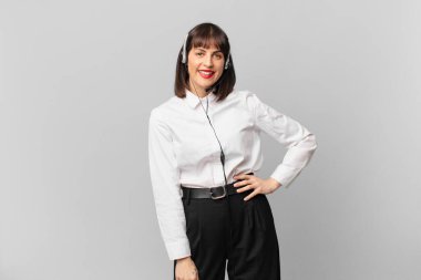 Tele pazarlamacı kadın mutlu bir şekilde gülümsüyor, eli kalçasında ve kendinden emin, olumlu, gururlu ve arkadaş canlısı.