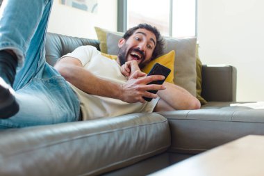 Genç sakallı adam akıllı telefonuyla kanepede dinleniyor.