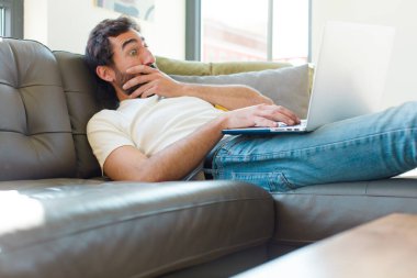 Genç sakallı bir adam dizüstü bilgisayarla kanepede dinleniyor.