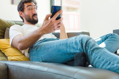 Genç sakallı adam akıllı bir telefonla kanepede dinleniyor.