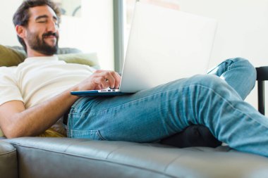 Genç sakallı bir adam dizüstü bilgisayarla kanepede dinleniyor.