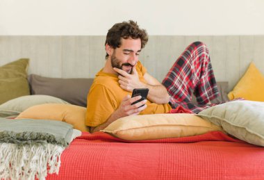 Yatakta cep telefonuyla sakallı genç bir adam.