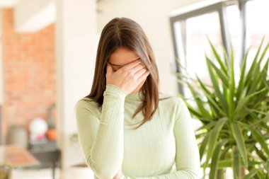 Güzel bir kadın stresli, utanmış ya da üzgün görünüyor, baş ağrısı var, yüzünü elleriyle kapatıyor.