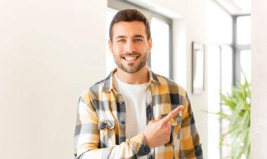 Yakışıklı adam neşeyle gülümsüyor, mutlu hissediyor ve yan ve yukarı doğru işaret ediyor, fotokopi uzayındaki nesneleri gösteriyor.