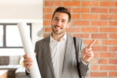 Yakışıklı mimar neşeyle gülümsüyor, mutlu hissediyor ve kenarı ve yukarıyı işaret ediyor, fotokopi uzayındaki nesneleri gösteriyor