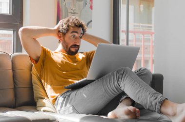 Genç sakallı bir adam stresli, endişeli, endişeli ya da korkmuş, elleri başının üstünde, bir dizüstü bilgisayarla yanlışlıkla panikliyor.