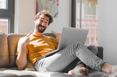 Genç sakallı adam kendi geniş gülümsemesine güvenerek gülümsüyor. Kanepe dizüstü bilgisayarındaki dizüstü bilgisayarla pozitif, rahat, tatmin olmuş bir tavır sergiliyor.