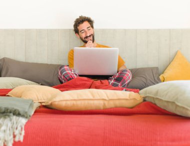 Genç sakallı bir adam mutlu, kendinden emin bir ifadeyle gülümsüyor. Elleri çenesinde, merak ediyor ve yan tarafa bakıyor.