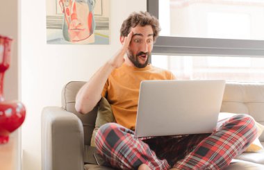 Genç sakallı adam mutlu, şaşkın ve şaşırmış görünüyor, gülümsüyor ve inanılmaz ve inanılmaz iyi haberleri fark ediyor ve dizüstü bilgisayarla oturuyor.