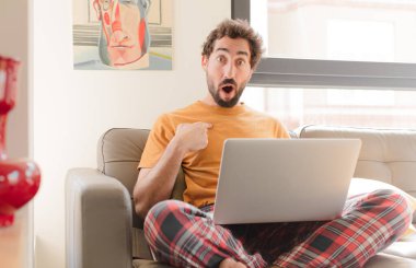 Genç sakallı adam şok olmuş ve şaşırmış görünüyor ağzı açık, kendini işaret ediyor ve dizüstü bilgisayarla oturuyor.