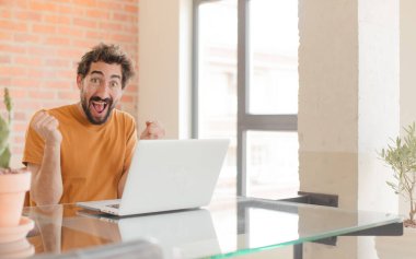Genç sakallı adam şok olmuş, heyecanlı ve mutlu hissediyor, gülüyor ve başarıyı kutluyor, vay canına! Ve bir dizüstü bilgisayarla çalışmak.
