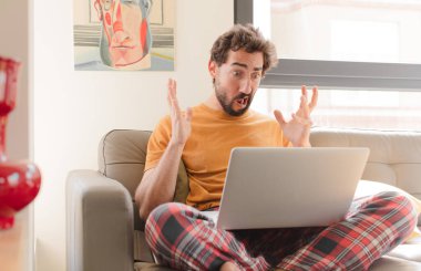 Genç sakallı adam elleri havada bağırıyor. Öfkeli, hayal kırıklığına uğramış, stresli, üzgün ve dizüstü bilgisayarla oturuyor.