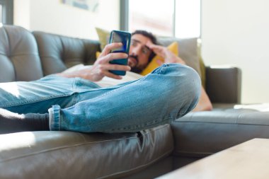 Genç sakallı adam akıllı telefonuyla kanepede dinleniyor.