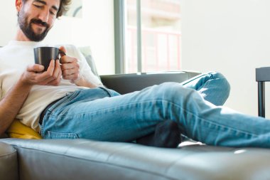 Genç sakallı adam elinde kahve fincanıyla kanepede dinleniyor.