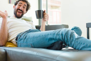 Genç sakallı adam elinde kahve fincanıyla kanepede dinleniyor.