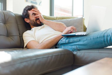 Genç sakallı bir adam dizüstü bilgisayarla kanepede dinleniyor.