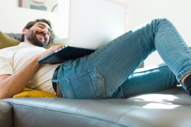 Genç sakallı bir adam dizüstü bilgisayarla kanepede dinleniyor.