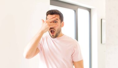 Yakışıklı adam şok olmuş, korkmuş ya da korkmuş görünüyor. Yüzü elle kaplanmış ve parmaklarının arasından gizlice bakıyor.