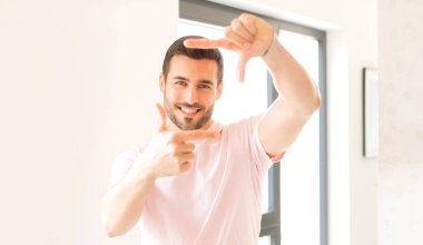 Yakışıklı adam mutlu, arkadaş canlısı ve pozitif hissediyor, gülümsüyor ve elleriyle bir portre veya fotoğraf çerçevesi yapıyor.