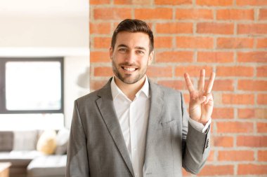 Yakışıklı iş adamı gülümsüyor ve arkadaş canlısı görünüyor. Üç ya da üç numarayı gösteriyor. Elleri önde, geriye doğru sayıyor.