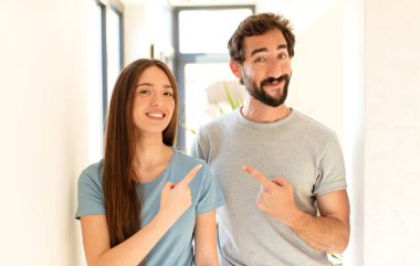 Genç çift neşeyle gülümsüyor, mutlu hissediyor ve kenarı ve yukarıyı işaret ediyor, fotokopi uzayındaki nesneleri gösteriyor