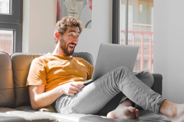 Genç sakallı adam mutlu ve hoş bir şekilde şaşırmış görünüyor. Kanepe dizüstü bilgisayarındaki dizüstü bilgisayarla büyülenmiş ve şok olmuş bir ifadeyle heyecanlanıyor.