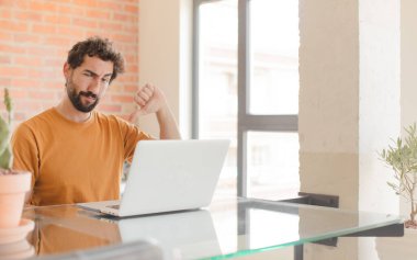 Genç sakallı adam sinirli, sinirli, hayal kırıklığına uğramış ya da canı sıkkın, ciddi bir bakışla baş parmağını kaldırıyor ve bir dizüstü bilgisayarla çalışıyor.
