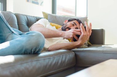 Genç sakallı adam akıllı telefonuyla kanepede dinleniyor.